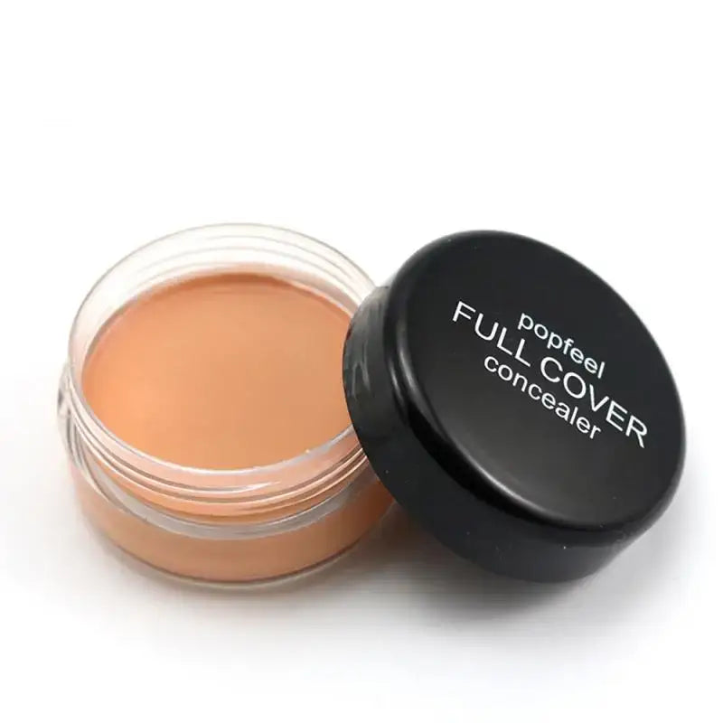 Open jar of cream-colored concealer makeup with a black lid labeled ’Full Cover concealer’.