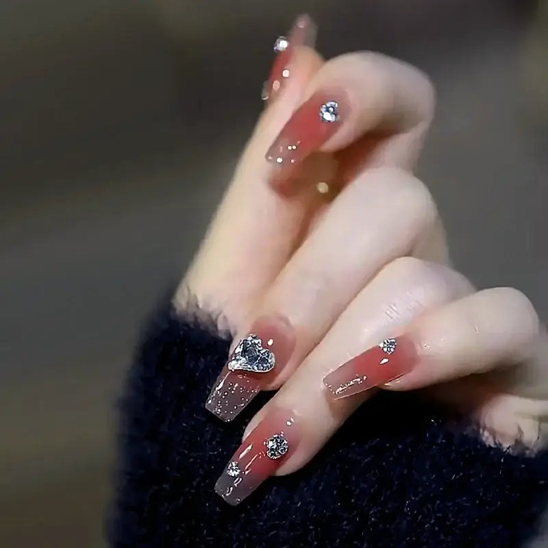 Manicured hand with long, decorated nails featuring red polish and rhinestone accents.