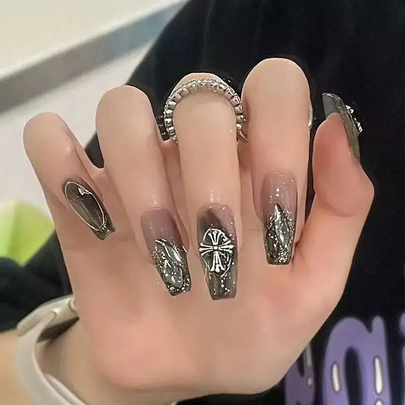 Hand with elaborately decorated long acrylic nails featuring metallic designs and a cross pattern.