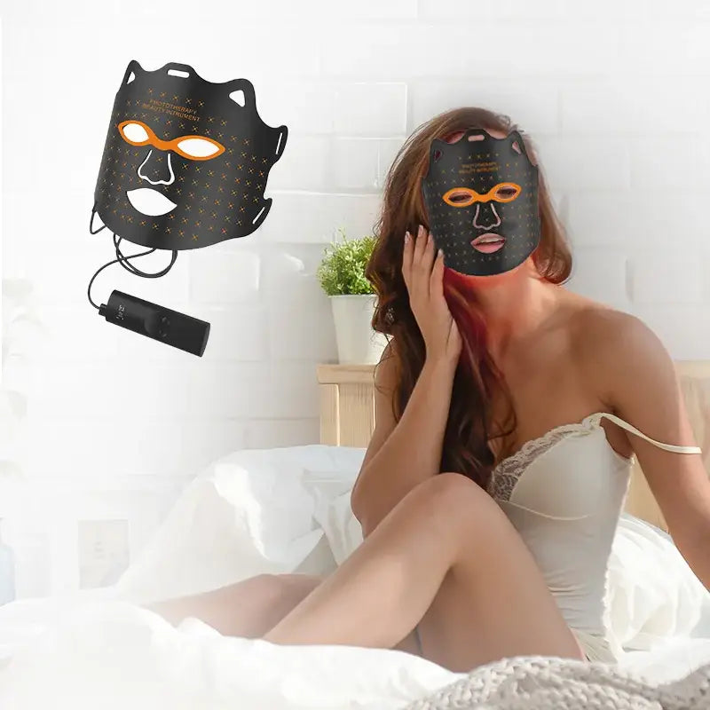 LED light therapy face mask worn by a person sitting on a bed.